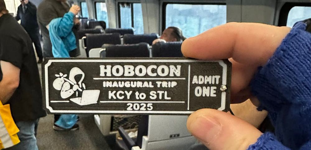 A 3D-printed badge reading HOBOCON, INAUGURAL TRIP, KCY to STL, 2025, ADMIT ONE, held up inside an Amtrak car