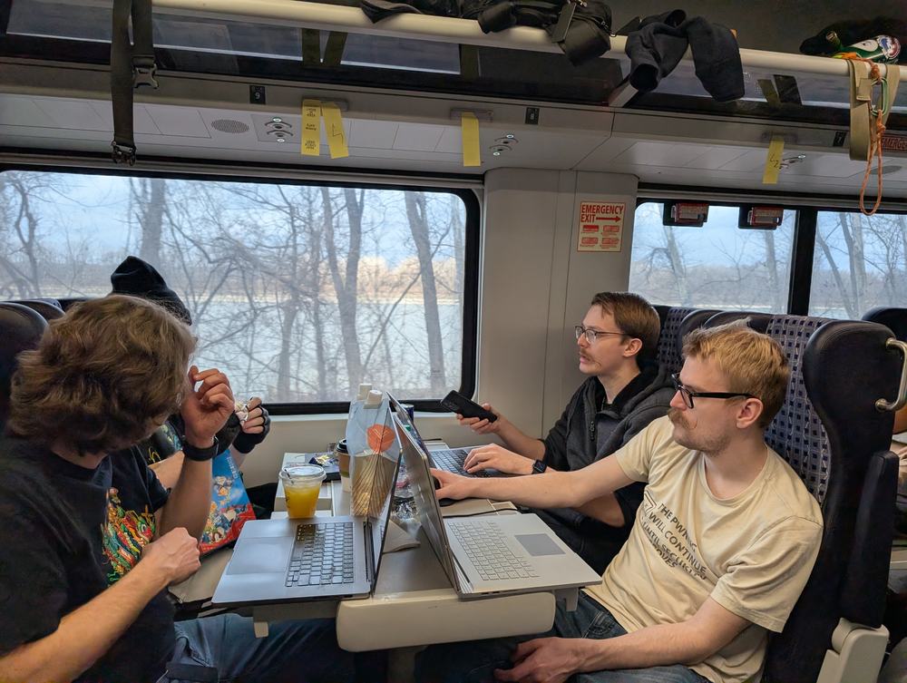 Four people working on laptops around a table on the return Amtrak train, trees and water visible out the window
