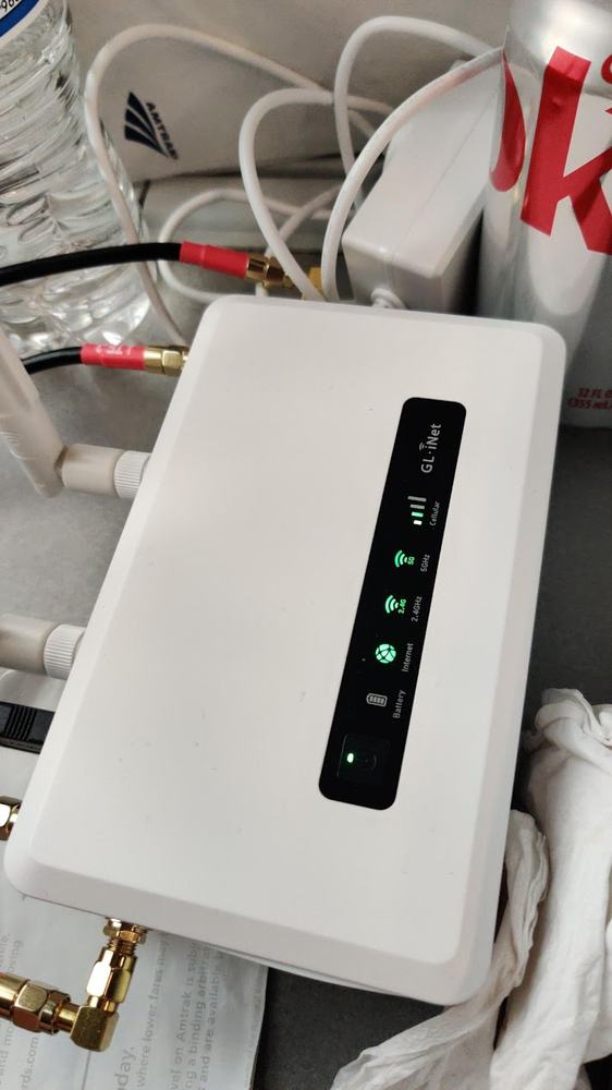 A GL.iNet travel router with external antennas attached, sitting on a train tray table among water bottles and cables, cellular and Wi-Fi status LEDs lit