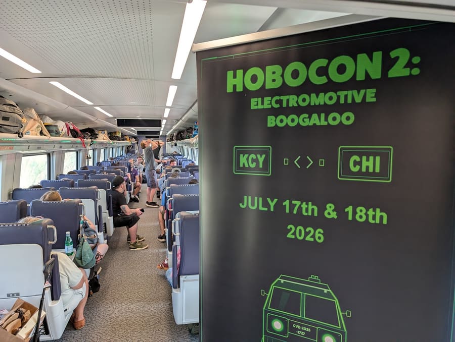 A tall banner standing in the aisle of the train car reading HOBOCON 2: ELECTROMOTIVE BOOGALOO, KCY to CHI, July 17th and 18th 2026, in green pixel lettering, with the packed car behind it
