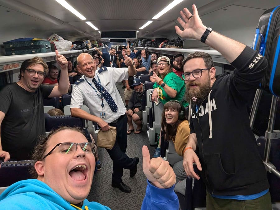 A wide selfie down the length of the train car with dozens of attendees cheering and giving thumbs up, the Amtrak cafe car attendant standing in the aisle among them holding a paper bag and laughing