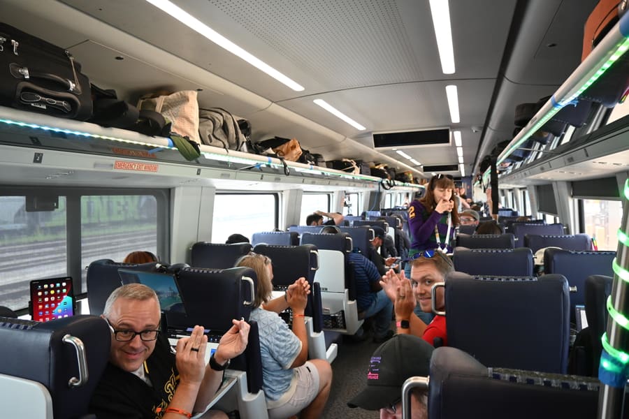 The interior of a packed Amtrak car full of Hobocon attendees, several turning to the camera and throwing hand signs, bags stuffed in the overhead racks and green LED strips running along them