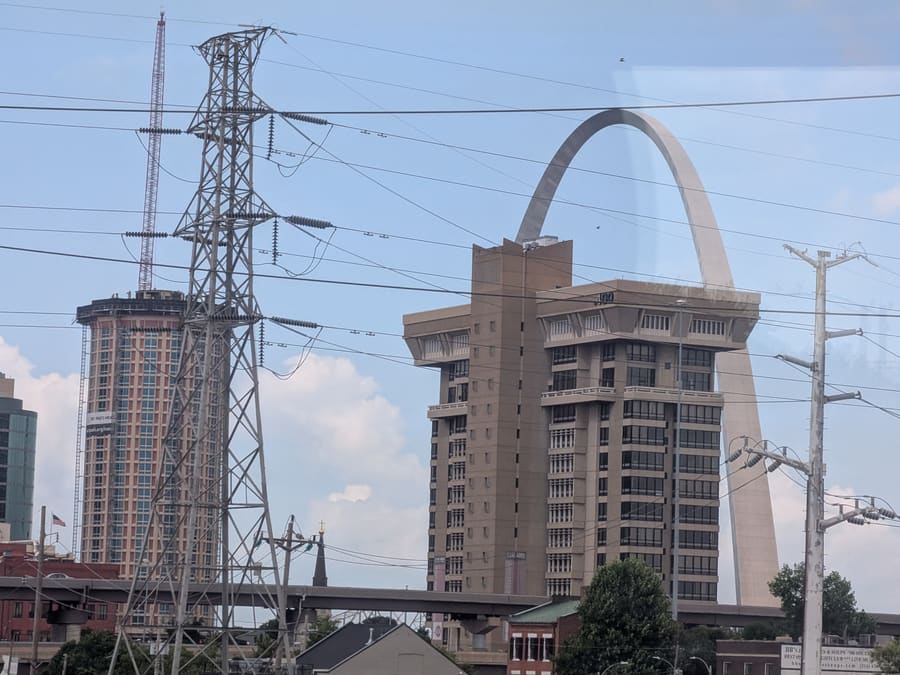 The Gateway Arch in St. Louis seen through a train window, framed between overhead catenary wires, a lattice transmission tower and downtown buildings