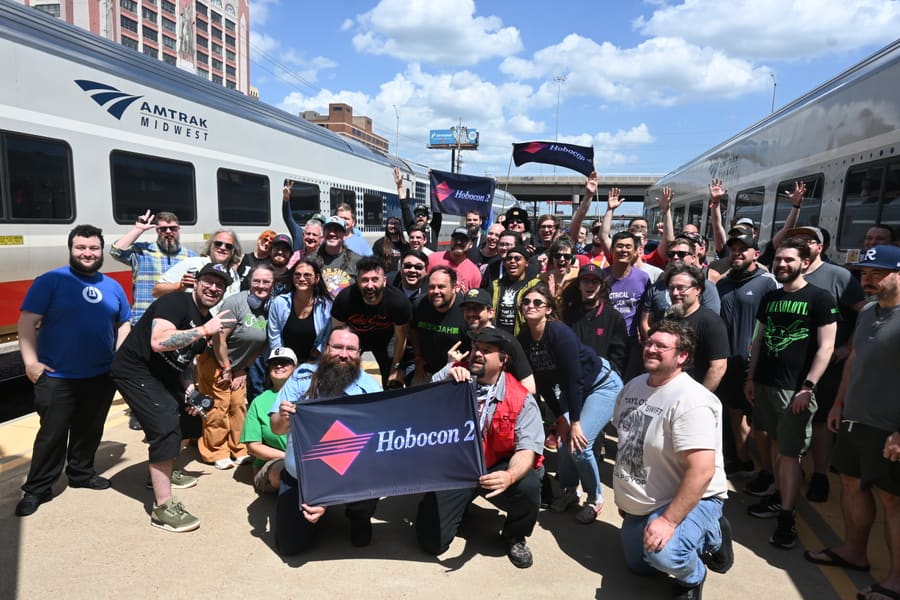 The full Hobocon 2 group posed on a sunny station platform between two Amtrak Midwest trainsets, holding a Hobocon 2 flag in front and waving two more overhead