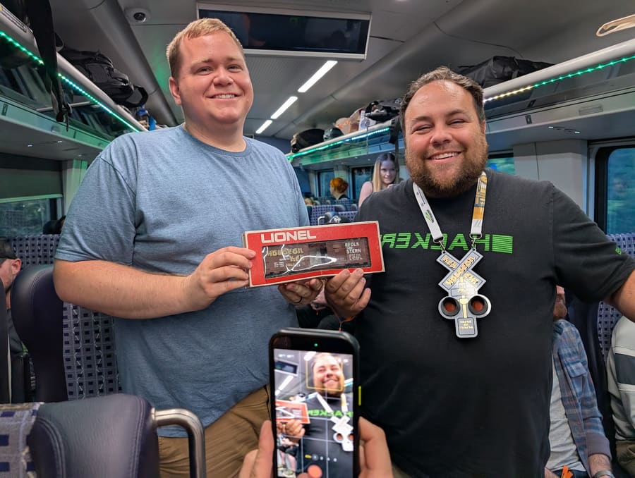 Two attendees in the train aisle, one holding up a boxed Lionel Norfolk and Western model boxcar hand-tagged in white paint pen, the other wearing a large 3D-printed railroad-crossing-signal badge on a lanyard, a phone filming them in the foreground