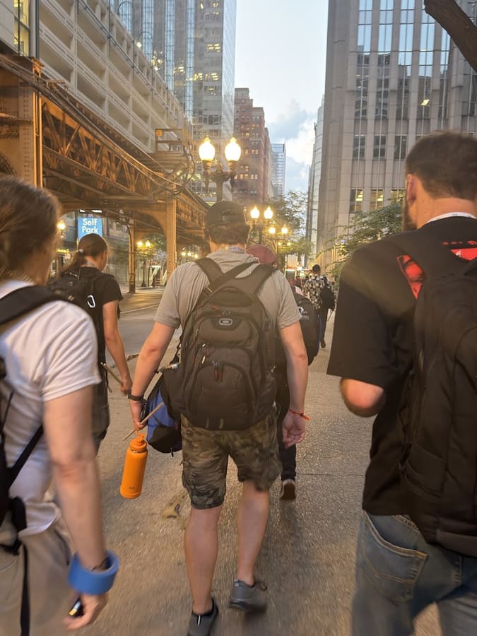 A group walking away from the camera along a Chicago sidewalk at golden hour, backpacks and bags on, elevated train tracks and lit street lamps ahead