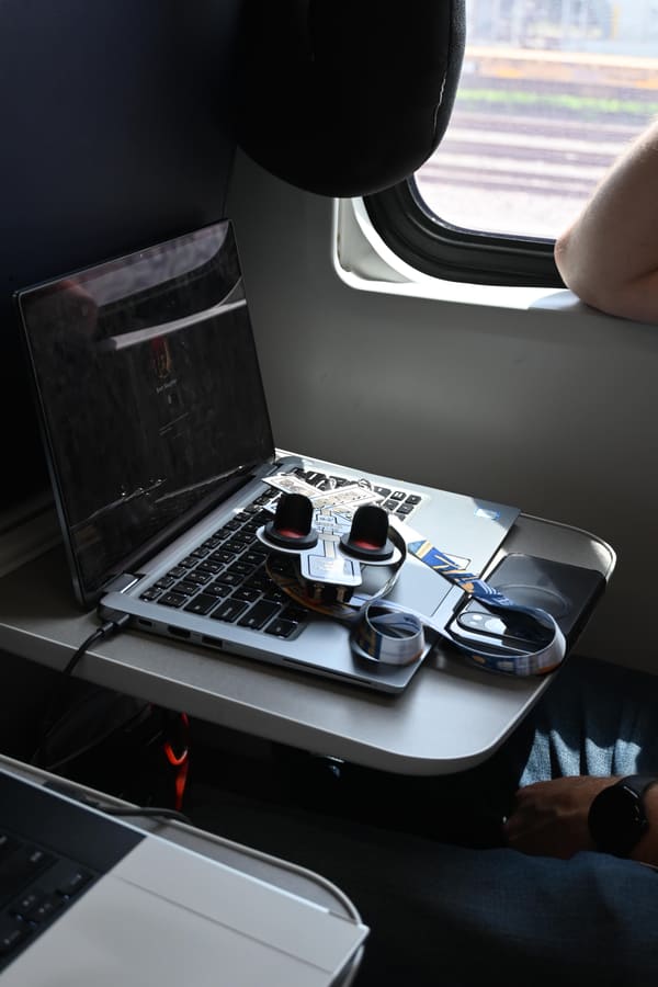 A sticker-covered laptop open on an Amtrak tray table with a small hardware board and knobs resting on the keyboard, window seat and daylight behind it