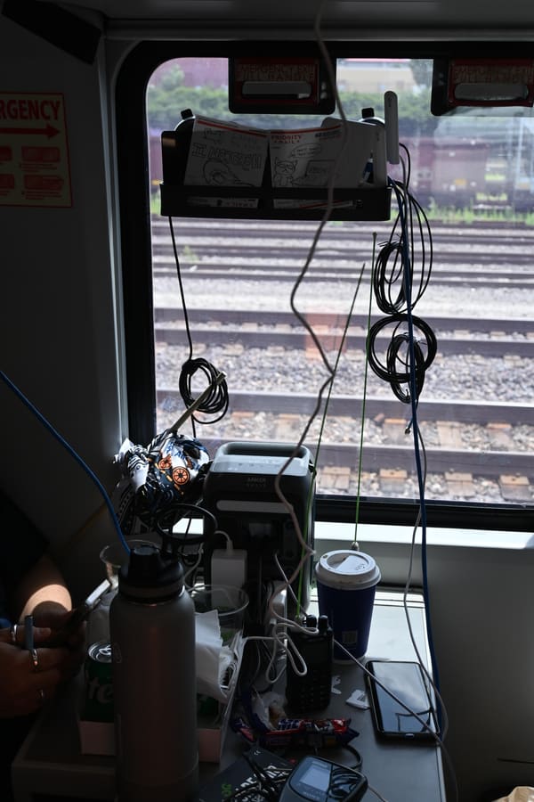 An improvised communications setup at a train window: antennas mounted in the glass, coils of coax and ethernet strung from the luggage rack, a handheld radio, phone and power bank on the tray table
